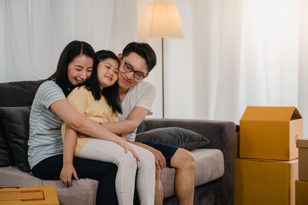 I giovani proprietari di case asiatici felici hanno comprato la nuova casa. Mamma, papà e figlia giapponesi che abbracciano in attesa del futuro nella nuova casa dopo essersi trasferiti nella delocalizzazione che si siede insieme sul sofà con le scatole.