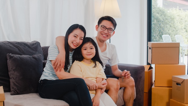 I giovani proprietari di case asiatici felici hanno comprato la nuova casa. Mamma, papà e figlia giapponesi che abbracciano in attesa del futuro nella nuova casa dopo essersi trasferiti nella delocalizzazione che si siede insieme sul sofà con le scatole.