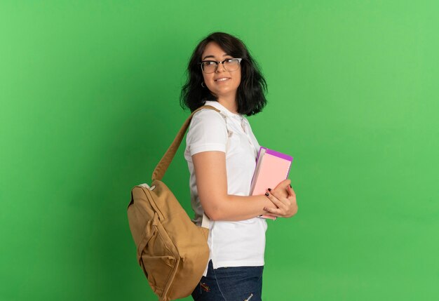 I giovani soddisfano la studentessa piuttosto caucasica con gli occhiali e la borsa posteriore si trovano lateralmente tenendo i libri guardando indietro sul verde con spazio di copia