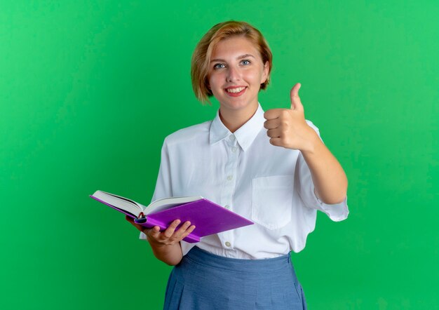 I giovani sorridenti bionda ragazza russa thumbs up tenendo il libro isolato su sfondo verde con copia spazio
