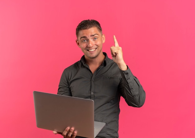 I giovani sorridenti biondi belli manholds laptop e punti in alto guardando la telecamera isolata su sfondo rosa con copia spazio