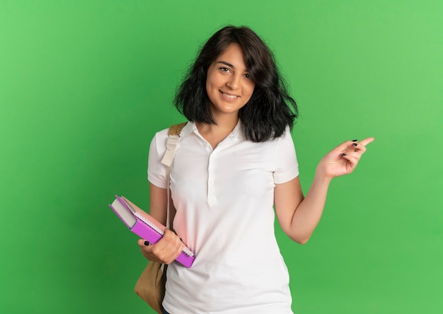 I giovani sorridenti studentessa piuttosto caucasica che indossa la borsa indietro punti a lato tenendo i libri su verde con copia spazio