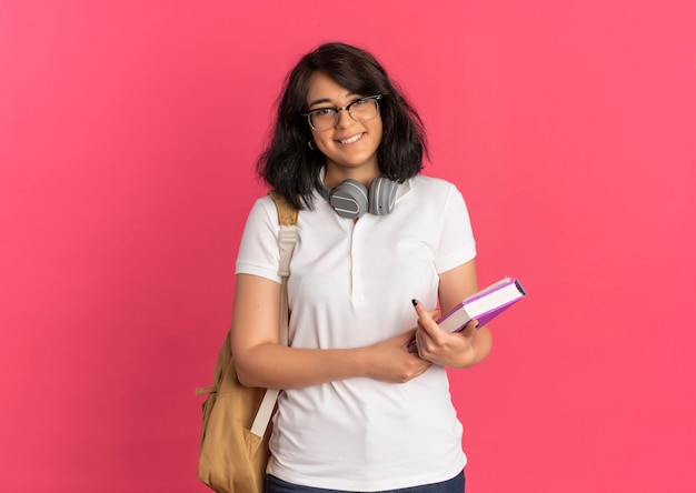 I giovani sorridenti studentessa piuttosto caucasica con gli occhiali cuffie e borsa posteriore tiene il libro sul rosa con spazio di copia