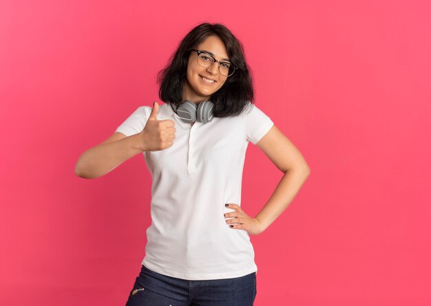 I giovani sorridenti studentessa piuttosto caucasica con gli occhiali e le cuffie sul collo pollici in su e mette la mano sulla vita in rosa con spazio di copia