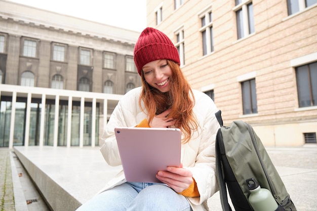 I sorrisi della ragazza rossa si siedono all'aperto vicino all'edificio con il thermos per tablet digitale e lo zaino a cui si collega