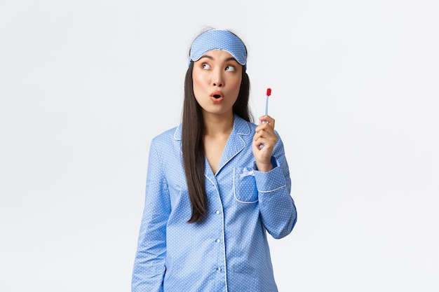 Igiene, stile di vita e concetto di persone a casa. La ragazza asiatica premurosa in pigiama e maschera per dormire ha una grande idea mentre si lava i denti, tiene lo spazzolino da denti e guarda l'angolo in alto a sinistra.