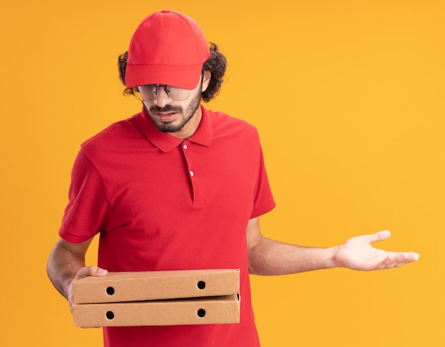 Ignaro giovane fattorino in uniforme rossa e berretto con gli occhiali che tengono in mano e guardano i pacchetti di pizza che mostrano la mano vuota isolata sul muro arancione