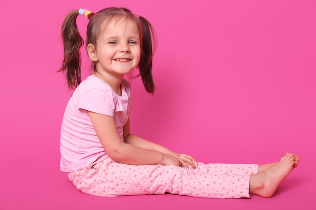 Il bambino abbastanza piccolo con due code di cavallo e molti scrunchies colorati si siede sul pavimento e felice di essere fotografato in studio fotografico. Adorabili sorrisi di bambini. Concetto di bambini e infanzia.