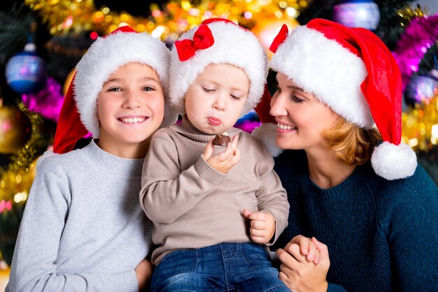 Il bambino mangia la caramella dolce in una cerchia di famiglia. Sorridente giovane madre con due figli in cappelli di Babbo Natale.