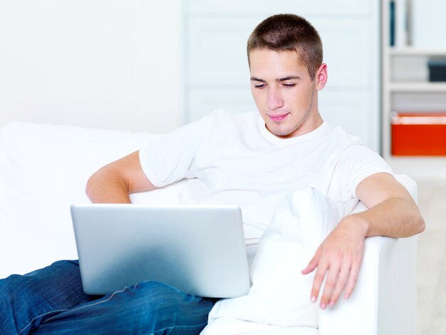 Il bel ragazzo giovane guarda nel laptop a casa