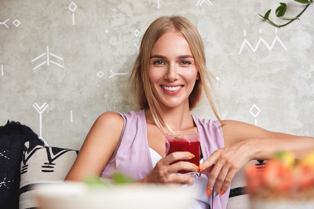 Il bello e giovane modello femminile positivo si sente rilassato mentre si siede sul comodo divano nella caffetteria, gode di un frullato di fragole fresche, ricrea durante le vacanze estive. Persone, riposo e concetto di stile di vita