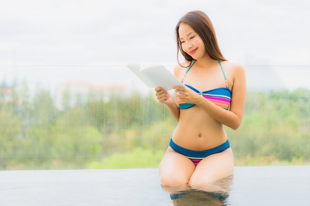 Il bello giovane sorriso asiatico della donna del ritratto felice si rilassa con il libro di lettura intorno alla piscina nella località di soggiorno dell&#39;hotel per svago