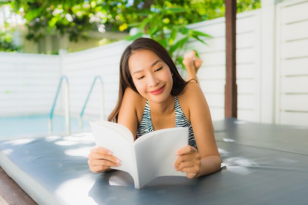 Il bello giovane sorriso asiatico della donna del ritratto felice si rilassa con il libro di lettura intorno alla piscina