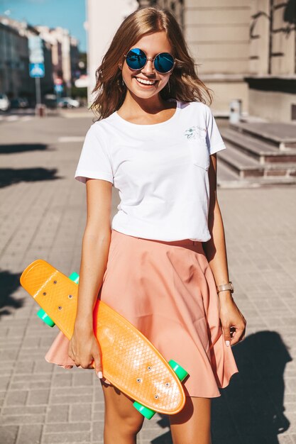 Il bello modello biondo sorridente sveglio dell'adolescente in pantaloni a vita bassa dell'estate copre con la posa arancio del pattino del penny