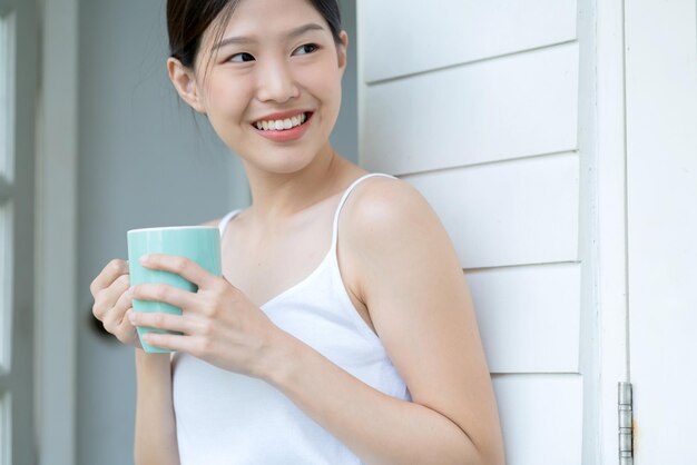 Il bello sorriso di felicità della canotta bianca asiatica teenager gode della freschezza del mattino con una bevanda calda vicino alla finestra con la luce del sole