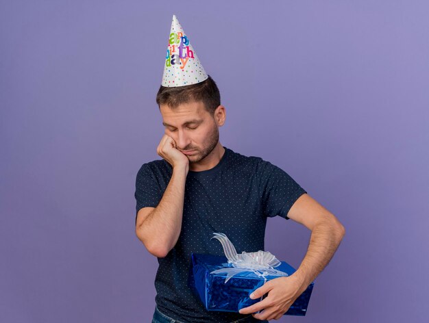 Il berretto da portare di compleanno dell'uomo bello triste mette la mano sul mento e tiene la confezione regalo isolata sulla parete viola con lo spazio della copia