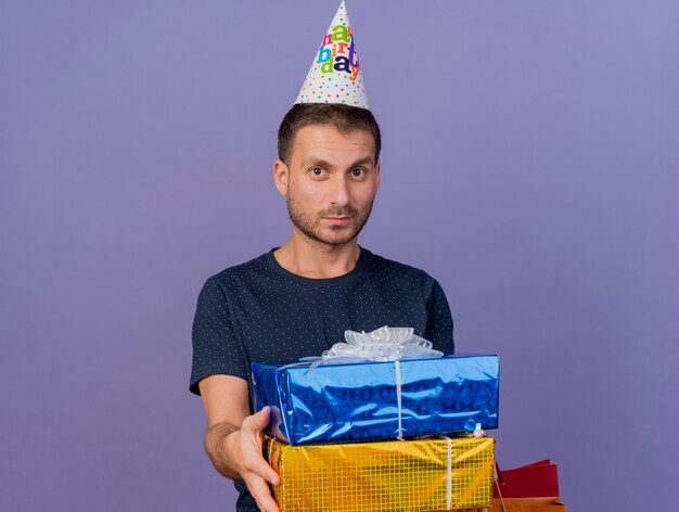 Il berretto da portare di compleanno dell'uomo caucasico bello soddisfatto tiene le scatole regalo e il sacchetto della spesa di carta isolato su fondo viola con lo spazio della copia