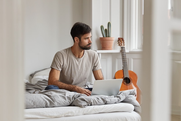 Il blogger maschio ha un'espressione premurosa, gode del tempo libero con il computer portatile, si siede su un letto comodo