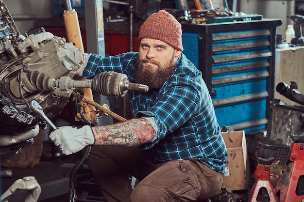 Il brutale meccanico barbuto tatuato ripara il motore dell'auto che viene sollevato sull'ascensore idraulico nel garage. Stazione di servizio.