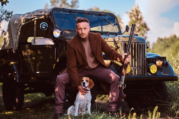 Il cacciatore in abiti eleganti tiene un fucile e si siede insieme al suo cane beagle mentre si appoggia a un'auto militare retrò in una foresta.