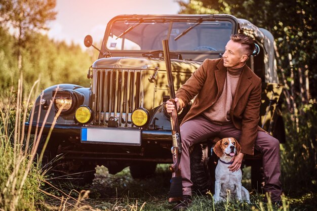 Il cacciatore in abiti eleganti tiene un fucile e si siede insieme al suo cane beagle mentre si appoggia a un'auto militare retrò in una foresta.