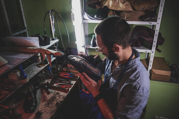 Il calzolaio diligente con gli occhiali sta lavorando con un paio di scarpe di pelle sul posto di lavoro.