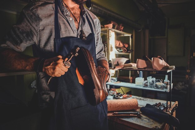 Il calzolaio diligente sta lavorando sulla suola delle scarpe usando uno strumento speciale nel suo posto di lavoro buio.