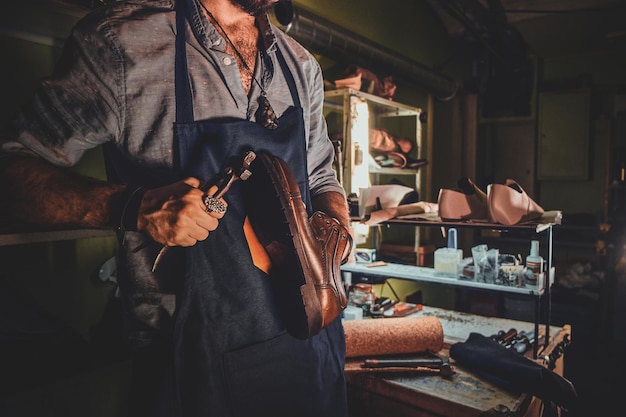 Il calzolaio diligente sta lavorando sulla suola delle scarpe usando uno strumento speciale nel suo posto di lavoro buio.