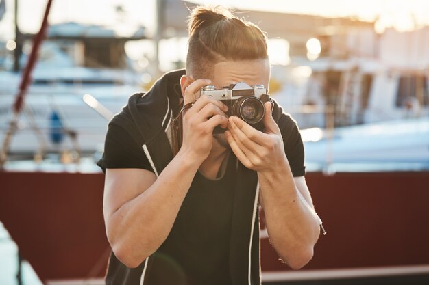 Il cameraman cerca di trattenere ancora di non spaventare gli uccelli. Ritratto di giovane fotografo maschio focalizzato guardando attraverso la fotocamera e accigliato, concentrandosi sul modello durante la sessione fotografica vicino al mare nel porto