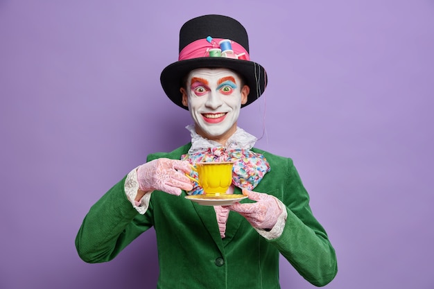 Il cappellaio pazzo positivo indossa un trucco colorato luminoso e si diverte a bere il tè alla festa vestita in costume e celebra le pose di Halloween felici contro il muro viola