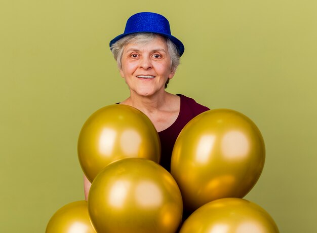 Il cappello da portare del partito della donna anziana sorridente sta con gli aerostati dell'elio isolati sulla parete verde oliva con lo spazio della copia