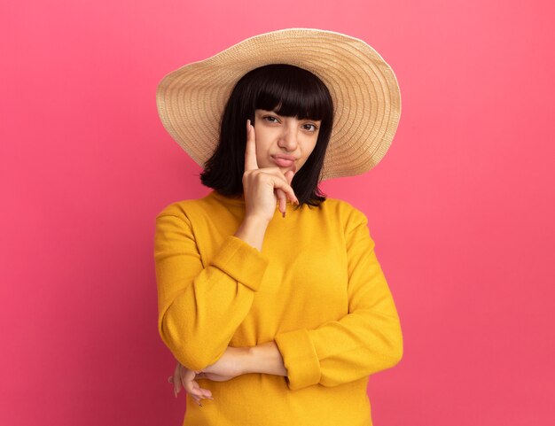 Il cappello da portare della spiaggia da portare della ragazza caucasica castana giovane insoddisfatta mette la mano sul mento e guarda la macchina fotografica sul rosa