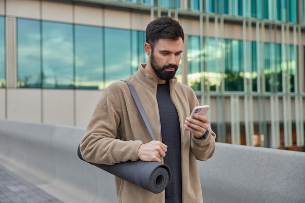 Il colpo all'aperto dell'uomo barbuto utilizza l'app per smartphone trasporta karemat arrotolato per fare pratica di yoga vestita in pose sportive contro esercizi di costruzione di vetro conduce attivamente uno stile di vita sano