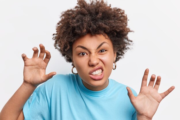 Il colpo alla testa della donna afroamericana fa ruggire o ringhiare il gesto delle zampe come un animale finge di essere un gatto stringe i denti indossa una maglietta blu casual isolata sul muro bianco. Espressioni facciali.