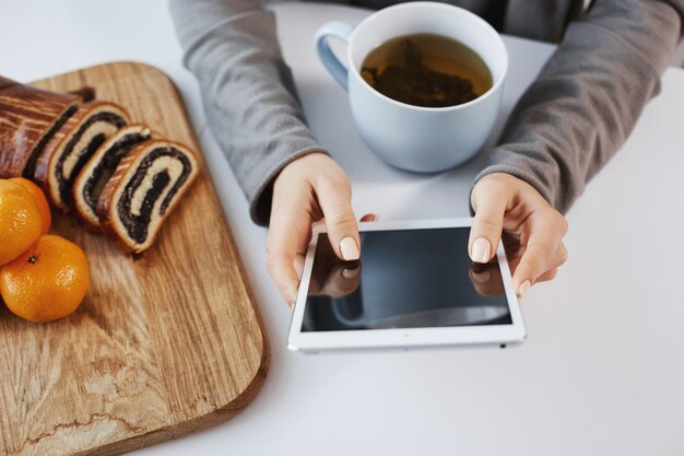 Il colpo del primo piano della donna passa la tenuta della compressa digitale. La ragazza ama i weekend in un'atmosfera calma e accogliente, bevendo una tazza di tè e mangiando mandarini con torta arrotolata. La donna di affari rimane sempre in contatto