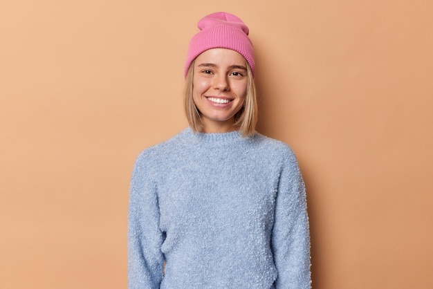 Il colpo di vita di una bella giovane donna sorride a denti stretti con un'espressione felice alla telecamera vestita con un cappello rosa casual jumper ha buon umore isolato su sfondo marrone. Concetto di emozioni positive