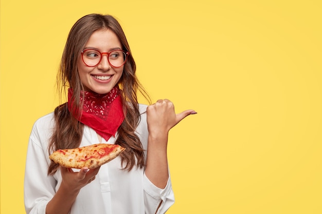 Il colpo isolato della ragazza sorridente attraente mostra la direzione alla pizzeria, mangia la pizza saporita con formaggio e pomodori, punta con il pollice allo spazio della copia contro il muro giallo. La donna ha uno spuntino al coperto