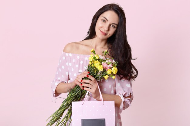 Il colpo isolato di attraente giovane donna europea ha i capelli lunghi neri, indossa un abito a pois, tiene un sacchetto regalo e fiori, pone sul muro rosa chiaro, celebra la giornata internazionale della donna