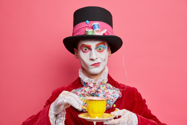 Il colpo orizzontale del cappellaio maschio serio pone con la tazza di tè indossa il cappello ha modi di abiti da gentiluomo aristocratici per le pose di carnevale mascherato al coperto