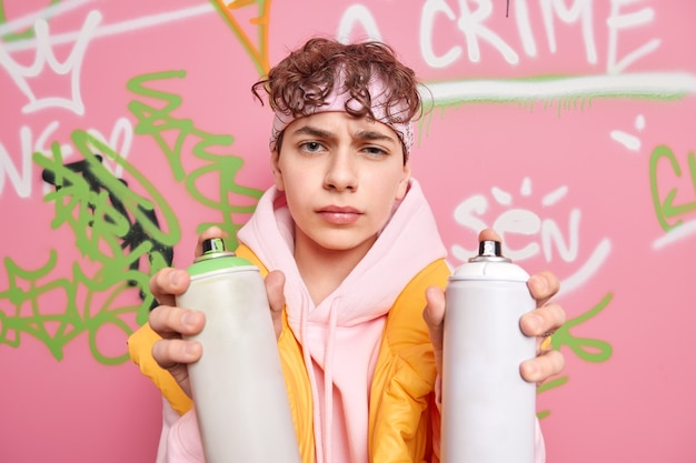 Il colpo orizzontale del ragazzo infelice giovane vestito in felpa tiene due bottiglie di aerosol fa graffiti creativi sul muro di strada ha un'espressione insoddisfatta