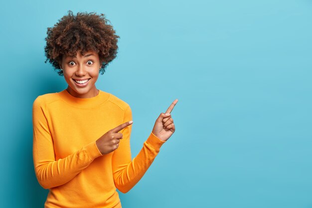 Il colpo orizzontale della bella donna dai capelli ricci indica lo spazio della copia della parete blu indica per la pubblicità e chiede non solo di camminare davanti ai sorrisi vestiti volentieri casualmente dà la giusta direzione