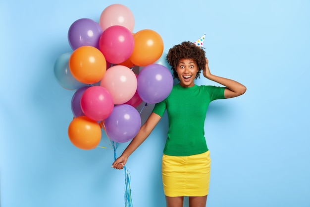 Il colpo orizzontale della donna felicissima tiene palloncini multicolori mentre posa con il cappello di compleanno