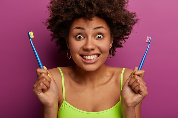 Il colpo ritagliato della donna dai capelli ricci ha un'espressione felice, tiene due spazzolini da denti, mostra i denti bianchi perfetti, ha una procedura igienica mattutina quotidiana, pelle sana, isolato sul muro viola.