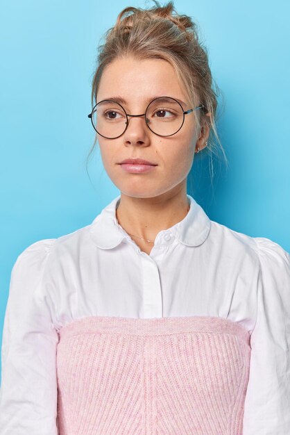 Il colpo verticale del modello femminile premuroso con i capelli pettinati concentrati lontano indossa pensosamente occhiali rotondi e vestiti puliti ha un'espressione intelligente isolata su sfondo blu considera qualcosa