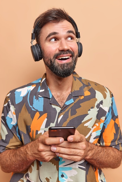 Il colpo verticale dell'uomo allegro barbuto tiene il telefono cellulare ascolta la musica tramite le cuffie senza fili