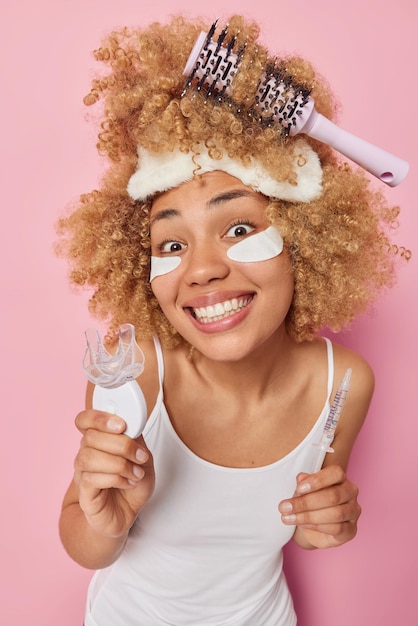 Il colpo verticale di una donna allegra tiene l'espansore della bocca in plastica che siringa con sorrisi liquidi vestito con i denti con una maglietta bianca casual si sottopone a trattamenti per la cura della pelle dopo il risveglio cerca di pettinare i capelli ricci