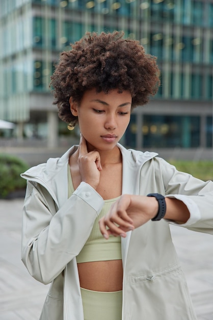 Il colpo verticale di una donna dai capelli ricci controlla il polso sul collo dopo che le attività fisiche guardano il moderno smartwatch indossa abiti sportivi all'aperto su sfondo sfocato Stile di vita sano