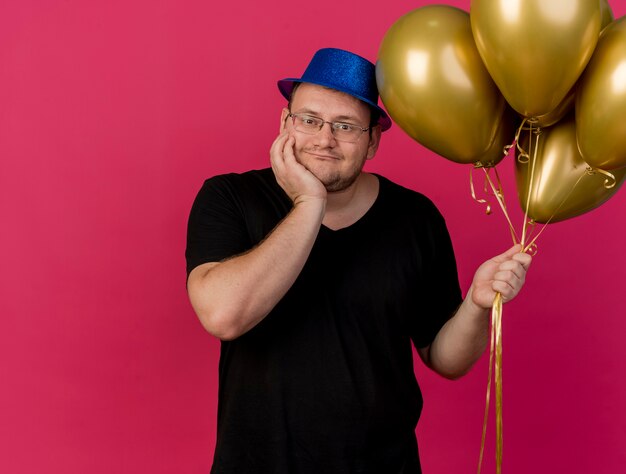 Il compiaciuto uomo slavo adulto in occhiali ottici che indossa un cappello da festa blu mette la mano sul mento e tiene palloncini di elio