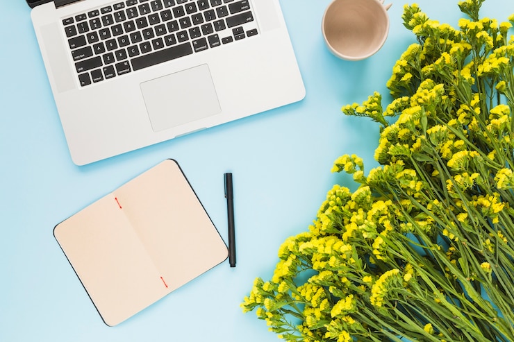 Foto gratuito il computer portatile; diario; penna; tazza di ceramica vuota e bouquet di fiori su sfondo blu