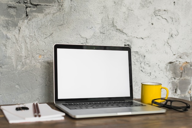 Il computer portatile; tazza; occhiali e blocco note sulla scrivania in legno